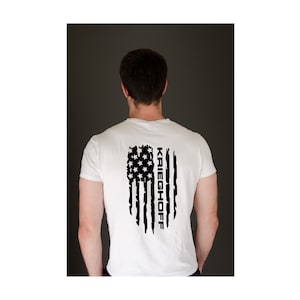 May include: White t-shirt with a black distressed American flag design and the name "KRIEGHOFF" printed vertically on the right side of the flag.