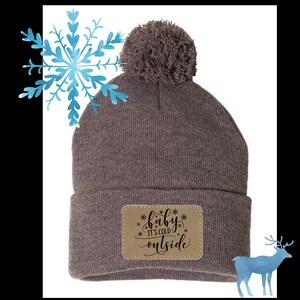Puede incluir: Gorro de punto gris con un parche de imitación de cuero marrón que dice "baby it's cold outside" con copos de nieve alrededor del texto. El gorro tiene un pompón esponjoso en la parte superior.