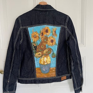 Può includere: Una giacca di jeans blu scuro con un dipinto di girasoli in un vaso sul retro. Il dipinto è in stile realistico e presenta girasoli gialli, foglie verdi e un vaso blu.