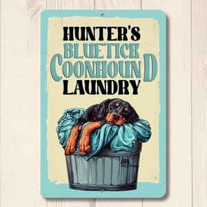 May include: A blue and white metal sign with the text "Hunter's Bluetick Coonhound Laundry" and a cartoon image of a black and brown Bluetick Coonhound dog sleeping in a laundry basket.