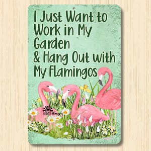 May include: Metal sign with a green background and white text that reads "I Just Want to Work in My Garden & Hang Out with My Flamingos". The sign features a watercolor illustration of three pink flamingos in a garden setting with yellow flowers and green grass.