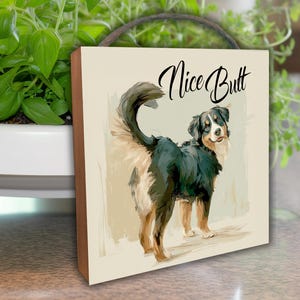 May include: A wooden wall art piece featuring a dog illustration with the text "Nice Butt" in a playful script. The dog is depicted with black, white, and tan fur, standing on a light background. The artwork has a rustic, warm aesthetic.