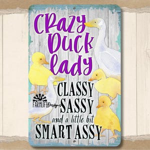 Puede incluir: Un letrero rectangular con borde azul claro y fondo de madera. Presenta un pato blanco y patitos amarillos, con el texto "Crazy Duck Lady, Classy, Sassy, and a little bit Smart Assy" en morado y negro.