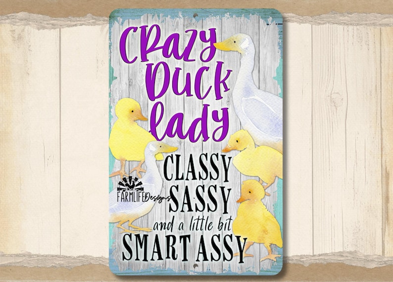 Puede incluir: Un letrero rectangular con borde azul claro y fondo de madera. Presenta un pato blanco y patitos amarillos, con el texto "Crazy Duck Lady, Classy, Sassy, and a little bit Smart Assy" en morado y negro.