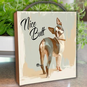 May include: A wooden wall art piece featuring a Boston Terrier dog with the text "Nice Butt" in a script font. The dog is depicted in shades of brown, white, and gray, with a collar. The background has a neutral color palette.