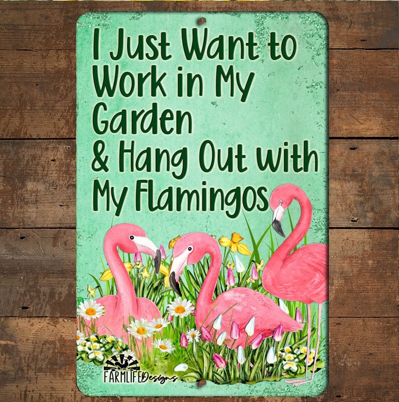 May include: A metal sign with a green background and white text that reads "I Just Want to Work in My Garden & Hang Out with My Flamingos". The sign features a pink flamingo with a yellow beak and a pink flamingo with a red beak. The flamingos are surrounded by green grass and flowers.