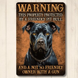 May include: A rectangular metal sign with a warning about property protection. The sign features a black pit bull illustration with text that reads "WARNING THIS PROPERTY PROTECTED BY A FRIENDLY PIT BULL AND A NOT SO FRIENDLY OWNER WITH A GUN."