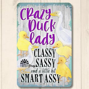 Puede incluir: Un letrero rectangular con borde azul claro y fondo de madera. Presenta un pato blanco y patitos amarillos, con el texto "Crazy Duck Lady, Classy, Sassy, and a little bit Smart Assy" en morado y negro.