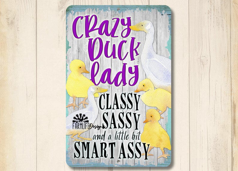 Puede incluir: Un letrero rectangular con borde azul claro y fondo de madera. Presenta un pato blanco y patitos amarillos, con el texto "Crazy Duck Lady, Classy, Sassy, and a little bit Smart Assy" en morado y negro.