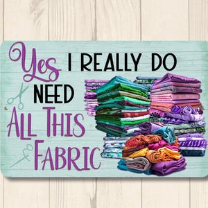 May include: A teal blue sign with the text "Yes I really do need all this fabric" in purple and black lettering. The sign features a pile of colorful fabric in the background.