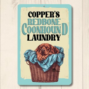 May include: A blue and white metal sign with the text "Copper's Redbone Coonhound Laundry" in a retro font. A brown and white Redbone Coonhound dog is sleeping in a wicker laundry basket.