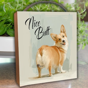 May include: A wooden wall art piece featuring a corgi dog illustration. The artwork has the words "Nice Butt" in a cursive font. The corgi is depicted in shades of brown and white, with a light blue background.