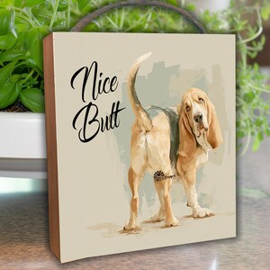 May include: Square wooden sign featuring a Basset Hound from the rear, with the text "Nice Butt" in elegant script. The dog is depicted in shades of tan, brown, and white, with long ears and a droopy face.