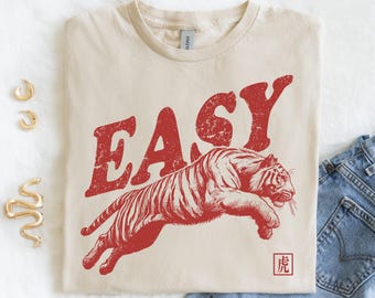 May include: Cream-colored t-shirt featuring a red graphic of a leaping tiger with the word "EASY" above it. The design includes a small Chinese character. The shirt is folded, with gold earrings and a gold snake ring visible.