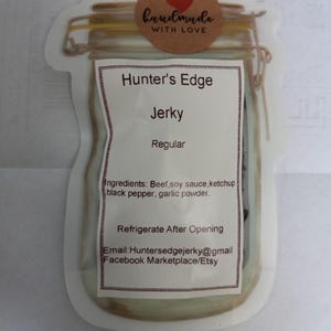 Op de afbeelding: Een witte sticker met een bruin potontwerp met de tekst "Hunter's Edge Jerky Regular". De sticker bevat ook de tekst "Ingrediënten: Rundvlees, sojasaus, ketchup, zwarte peper, knoflookpoeder." en "Na opening in de koelkast bewaren". De sticker bevat ook contactgegevens: "Email: Huntersedgejerky@gmail Facebook Marketplace/Etsy".