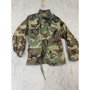May include: Camouflage pattern jacket with multiple pockets and a button-up front. The jacket features a stand-up collar and a drawstring at the waist. The color scheme includes shades of green, brown, and black, creating a military-inspired aesthetic.