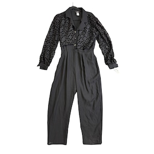 Puede incluir: Mono negro con una parte superior con cuello y mangas largas con estampado de lunares. El mono tiene una parte delantera abotonada y pantalones de pernera ancha. El conjunto está colgado de una percha negra.