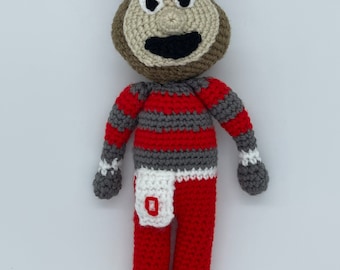 May include: Crocheted plush toy of a mascot wearing a red and white hat with a grey and red striped shirt and red pants with a white stripe and a white pocket with a red "O" on it.
