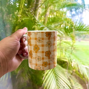May include: A white ceramic mug with a yellow and orange checkered pattern and floral design. The mug is being held by a hand.