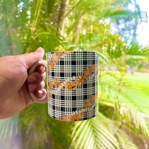 May include: A white ceramic mug with a black and white plaid pattern and a floral design in orange and white. The mug has a white handle.