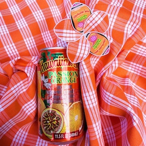May include: An 11.5 fl oz aluminum can of Hawaiian Sun Passion Orange juice drink featuring a graphic of a passion fruit and an orange. The can is adorned with a decorative flower-shaped cutout made of orange and white plaid fabric.