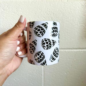 May include: White ceramic mug featuring a repeating pattern of black and white abstract shapes. The mug has a white handle and the text "@CHAYBEES" is printed on the side. The mug is held by a person.