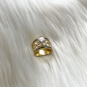 May include: A gold ring with a large clear stone set in the center. The band is decorated with a delicate floral design.
