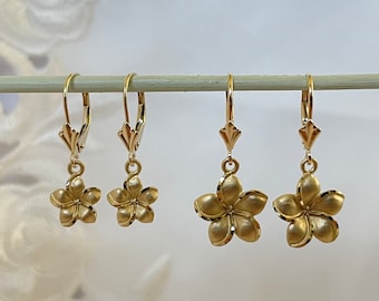 May include: Four pairs of gold-colored flower earrings. The earrings are lever-back style with a matte finish. The flower design has multiple petals. The earrings are displayed on a light-colored wooden dowel against a soft, white background.