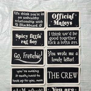 Peut inclure: Plusieurs écussons noirs et blancs avec des phrases amusantes. On peut lire des phrases comme « Official Mateys », « Spicy little rat boy » et « The Crew ».