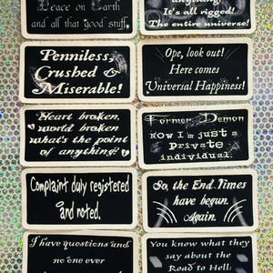 May include: A set of ten rectangular black fabric patches with white embroidered text. Each patch features unique phrases including Joy Unbounded, You can't change anything, Penniless, Crushed & Miserable, Ope, look out, Heart broken, Former Demon, Complaint duly registered, So, the End Times have begun, I have questions, and Road to Hell.