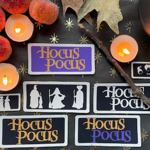 May include: A collection of Hocus Pocus-themed patches and candles arranged on a black surface with gold stars. The patches feature the movie title and silhouette designs of the Sanderson sisters. The candles are lit, creating a warm, inviting atmosphere.