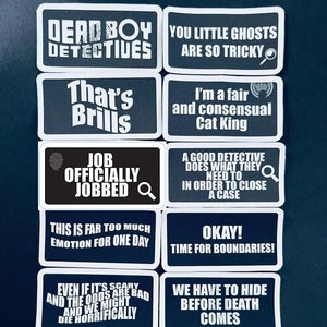 May include: A collection of black and white rectangular patches with various phrases in white text. Phrases include "Dead Boy Detectives," "You Little Ghosts Are So Tricky," and "Job Officially Jobbed."