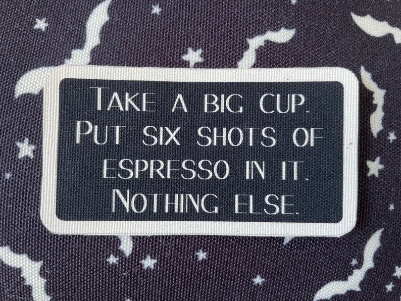 May include: A rectangular black and white sign with rounded corners. The sign reads "TAKE A BIG CUP. PUT SIX SHOTS OF ESPRESSO IN IT. NOTHING ELSE." The background is a dark fabric with white bats and stars.