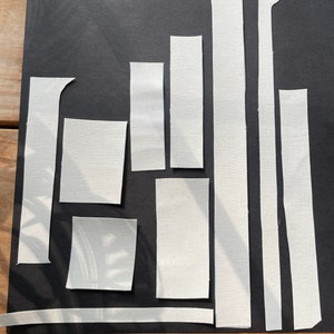 May include: Arrangement of white canvas fabric pieces on a black background. The pieces vary in shape and size, including rectangles and strips. The fabric has a textured appearance, and the composition is lit by natural light.