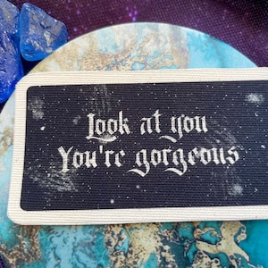 May include: A black rectangular patch with the words "Look at you, You're gorgeous" in white, ornate lettering. The patch has a white border and is on a blue and white marbled surface. Two blue crystals are in the upper left corner.