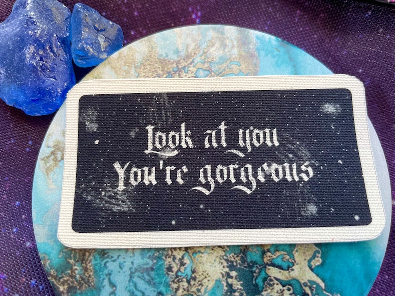 May include: A black rectangular patch with the words "Look at you, You're gorgeous" in white, ornate lettering. The patch has a white border and is on a blue and white marbled surface. Two blue crystals are in the upper left corner.