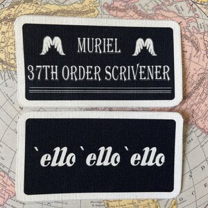 May include: Two rectangular black patches with white text and borders. The top patch reads "MURIEL 37TH ORDER SCRIVENER" with wing-like symbols. The bottom patch says "'ello 'ello 'ello". The patches are on a vintage map.