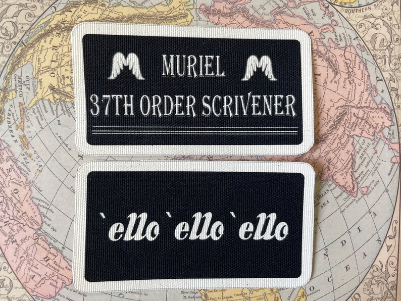 May include: Two rectangular black patches with white text and borders. The top patch reads "MURIEL 37TH ORDER SCRIVENER" with wing-like symbols. The bottom patch says "'ello 'ello 'ello". The patches are on a vintage map.