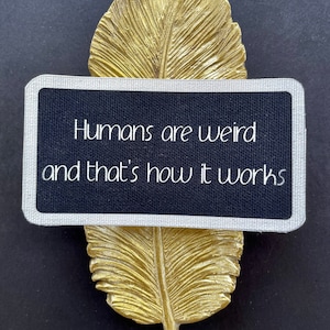 May include: A gold-coloured feather ornament with a rectangular black sign. The sign has a silver border and the text "Humans are weird and that's how it works" in white. The background is black.