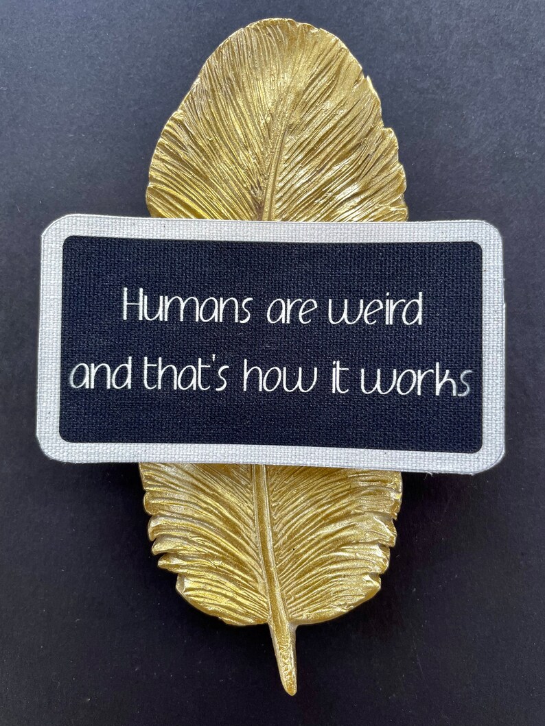May include: A gold-coloured feather ornament with a rectangular black sign. The sign has a silver border and the text "Humans are weird and that's how it works" in white. The background is black.