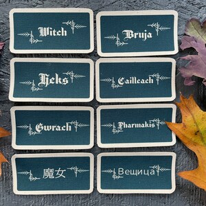 May include: Eight rectangular teal cards with white text, including the words "Witch," "Bruja," "Heks," "Cailleach," "Gwrach," "Pharmakis," "魔女," and "Вещица." The cards are arranged on a dark surface with colorful autumn leaves.