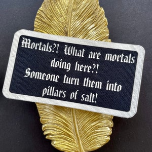 May include: A gold-coloured feather-shaped decorative item with a black rectangular card. The card has white text that reads "Mortals?! What are mortals doing here?! Someone turn them into pillars of salt!"