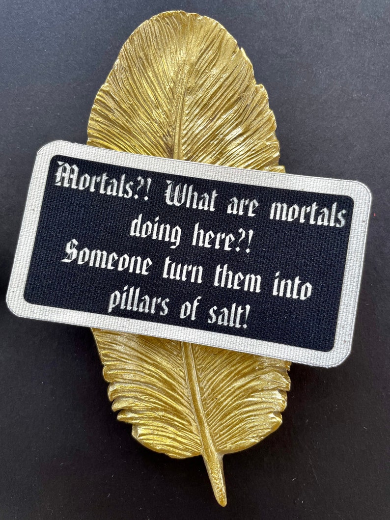 May include: A gold-coloured feather-shaped decorative item with a black rectangular card. The card has white text that reads "Mortals?! What are mortals doing here?! Someone turn them into pillars of salt!"