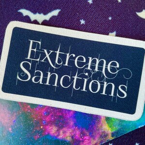 May include: A navy blue rectangular card with the words "Extreme Sanctions" in white script. The card has a white border and sits on a card with a colourful nebula design. The background is a dark purple fabric with white bat and star patterns.