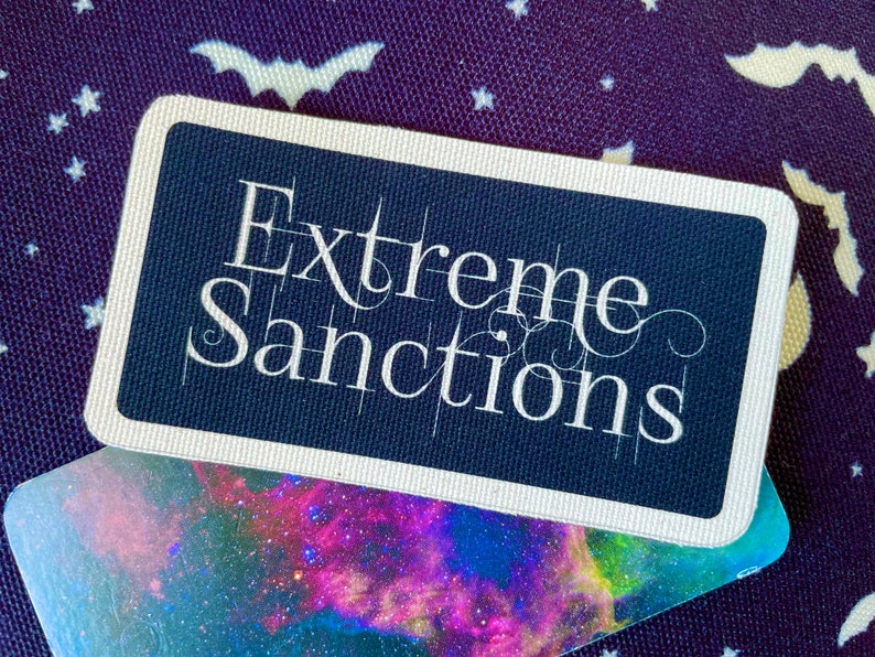 May include: A navy blue rectangular card with the words "Extreme Sanctions" in white script. The card has a white border and sits on a card with a colourful nebula design. The background is a dark purple fabric with white bat and star patterns.