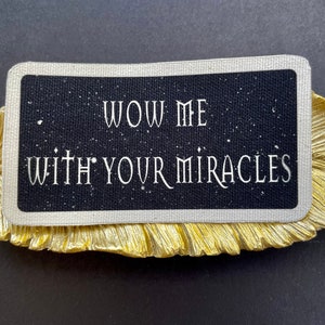 May include: A black card with white text reading "WOW ME WITH YOUR MIRACLES" sits on a gold-coloured, leaf-shaped decorative dish. The card has a starry background and a silver border. The dish is on a black surface.