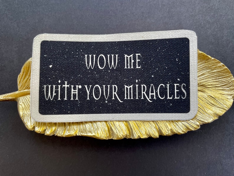 May include: A black card with white text reading "WOW ME WITH YOUR MIRACLES" sits on a gold-coloured, leaf-shaped decorative dish. The card has a starry background and a silver border. The dish is on a black surface.