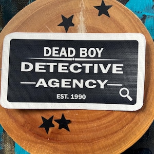 May include: A rectangular black and white patch with the text "DEAD BOY DETECTIVE AGENCY" and "EST. 1990". The patch has a white border and a magnifying glass icon. The patch is on a wooden surface with black star shapes.