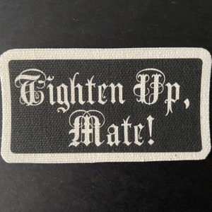 Pode incluir: Um patch retangular preto com uma borda branca. O patch apresenta a frase "Tighten Up, Mate!" em uma fonte gótica. O texto branco contrasta com o fundo preto, criando um efeito marcante.