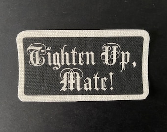 May include: A black rectangular patch with a white border. The patch displays the phrase "Tighten Up, Mate!" in a gothic font. The white text stands out against the black background, creating a striking contrast.